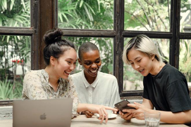 Drie mensen zitten rond een tafel in een café achter een laptop, lachend en wijzend naar een mobiele telefoon. Afbeelding van Ketut Subiyanto op Pexels.